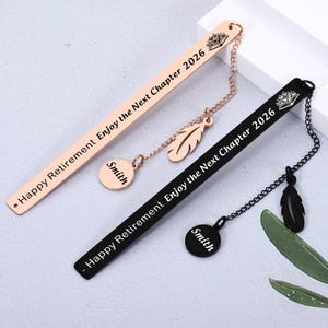 Pode incluir: Dois marcadores de metal, um em ouro rosa e outro preto, com o texto "Happy Retirement Enjoy the Next Chapter 2026". Cada um tem um pingente de pena e uma etiqueta redonda com "Smith". O marcador em ouro rosa tem um design de coroa.