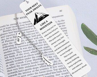 May include: A white bookmark with the text "YOUR NEXT PEAK AWAITS" and "LIKE A MOUNTAIN" with a mountain graphic. It includes inspirational text and a silver chain with a feather charm and a name tag that says "Leo".
