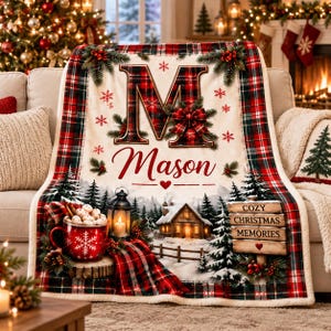 Könnte beinhalten: Eine cremefarbene Fleecedecke mit einem großen Buchstaben M, dem Namen Mason und einem rot-grünen Karomuster am Rand. Das Design zeigt eine rote Tasse mit Marshmallows, eine Laterne, eine Hütte im Schnee und ein Schild mit COZY CHRISTMAS MEMORIES.