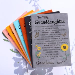 May include: A stack of notebooks in various colors, including pink, yellow, blue, orange, and black. The front notebook is gray and features the text "To My Granddaughter" with a floral and butterfly design. The notebooks are a thoughtful gift.