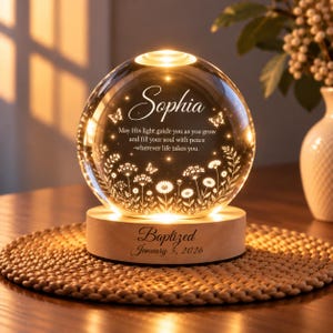 May include: A spherical glass night light on a round wooden base. The glass features an engraved floral design, butterflies, and the text: Sophia, May His light guide you as you grow and fill your soul with peace -wherever life takes you. The base reads: Baptized January 5, 2026.