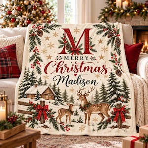 May include: A cream-coloured throw blanket featuring a large red letter M, the text Merry Christmas Madison, a snowy landscape with a cabin, a deer, and a fawn, decorated with pine branches, red berries, pinecones, and gold snowflakes.