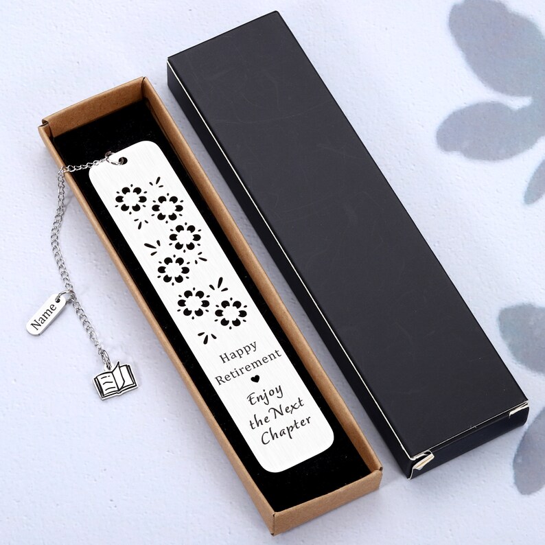 Puede incluir: Un marcap&aacute;ginas plateado con una cadena y un peque&ntilde;o encanto de libro. El marcap&aacute;ginas tiene un dise&ntilde;o recortado de fuegos artificiales y el texto "Happy Retirement Enjoy the Next Chapter".