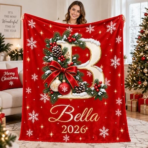 May include: A red fleece blanket featuring a large white letter B decorated with pine branches, red berries, pinecones, and a red bow. The blanket includes the name Bella and the year 2026 in gold script, surrounded by white snowflake patterns.