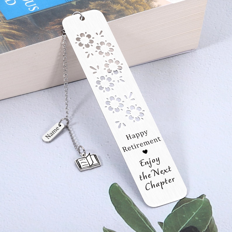 Puede incluir: Marcap&aacute;ginas de plata con un dise&ntilde;o recortado de fuegos artificiales y el texto "Happy Retirement Enjoy the Next Chapter". El marcap&aacute;ginas tiene un peque&ntilde;o amuleto de plata unido a una cadena que dice "Name" y un peque&ntilde;o amuleto de plata de un libro abierto.