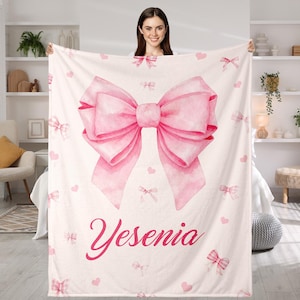 Puede incluir: Una manta rosa claro con un gran lazo de acuarela en el centro, rodeado de lazos y corazones más pequeños. El nombre "Yesenia" está escrito en una fuente rosa a juego debajo del lazo. La manta está sostenida por una persona.