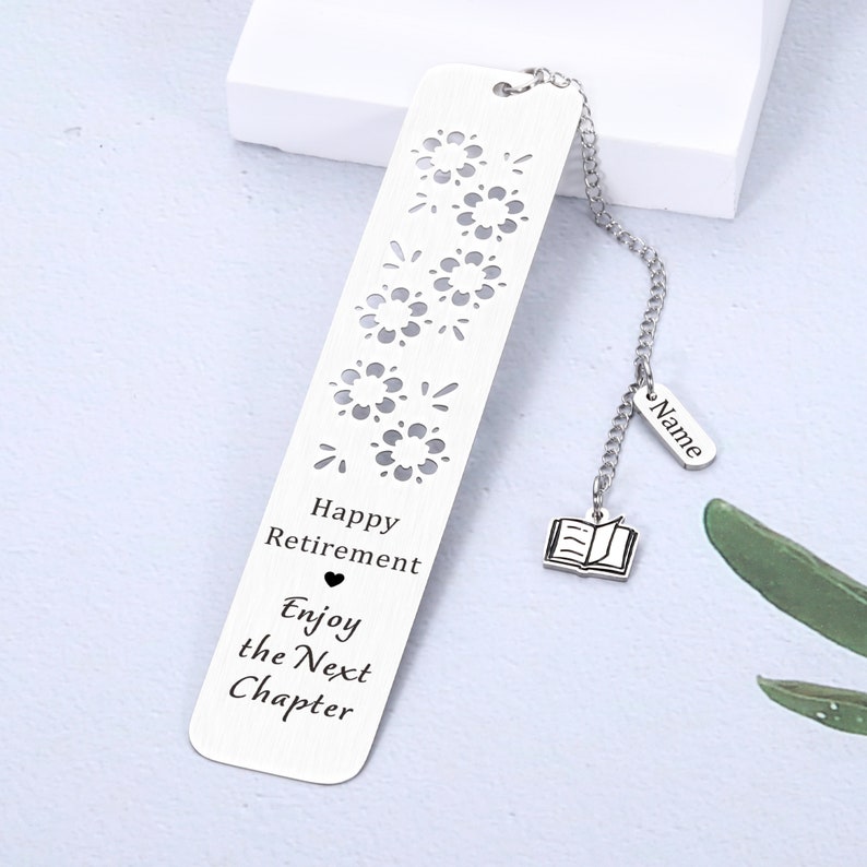 Puede incluir: Marcap&aacute;ginas de plata con una cadena y un peque&ntilde;o encanto de libro. El marcap&aacute;ginas tiene un dise&ntilde;o recortado de fuegos artificiales y el texto "Happy Retirement Enjoy the Next Chapter".
