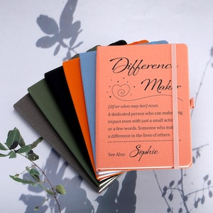 May include: A selection of notebooks in shades of grey, green, black, orange, and blue. The top notebook is coral and features the words "Difference Maker" and a definition. A pink elastic band secures the notebook.