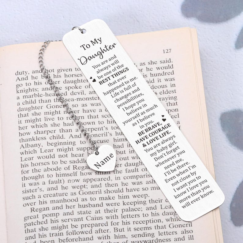 May include: Silver bookmark with a heart charm and a message that reads "To My Daughter" and includes a quote about being brave, having courage, and loving life. The bookmark is on a book with a brown cover.