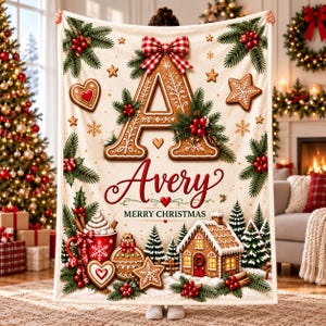 Op de afbeelding: Een crèmekleurige plaid met een grote peperkoekletter A bekroond met een rood-wit geblokte strik. Het ontwerp bevat de naam Avery, de tekst Merry Christmas, peperkoekkoekjes, dennentakken, rode bessen en een huisje.