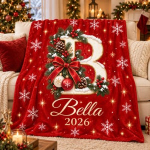 Op de afbeelding: Een rode fleece deken met een grote witte letter B versierd met dennentakken, rode bessen, dennenappels en een rode strik. De deken toont witte sneeuwvlokken, de naam Bella en het jaartal 2026 in een elegant goudkleurig lettertype.