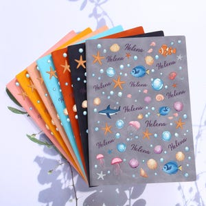 Pode incluir: Uma coleção de cadernos coloridos com tema marinho. Os cadernos apresentam ilustrações de estrelas do mar, conchas, peixes, águas-vivas e bolhas. O nome "Helena" é impresso em cada capa.