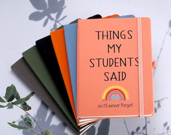 Pode incluir: Uma pilha de cadernos em várias cores, incluindo cinza, verde, preto, laranja e azul. O caderno da frente é cor de pêssego e apresenta o texto "THINGS MY STUDENTS SAID" com um gráfico de arco-íris e a frase "so I'll never forget."