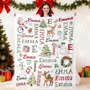 Peut inclure: Une couverture blanche sur le thème de Noël avec le nom "Emma" en rouge et noir, ornée d'illustrations festives du Père Noël, de rennes, de sapins et de bonhommes de neige. La couverture est présentée par une personne.