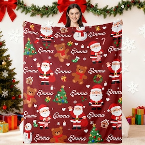 Puede incluir: Una manta navideña burdeos con diseños de Papá Noel, osos de peluche, árboles de Navidad y copos de nieve. El nombre "Emma" está repetido. La manta es sostenida por una persona.