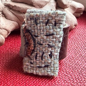 May include: A handcrafted ring with a woven fabric design, black stitching, and a brown leather band. The ring has a textured appearance with neutral colours and a touch of brown.