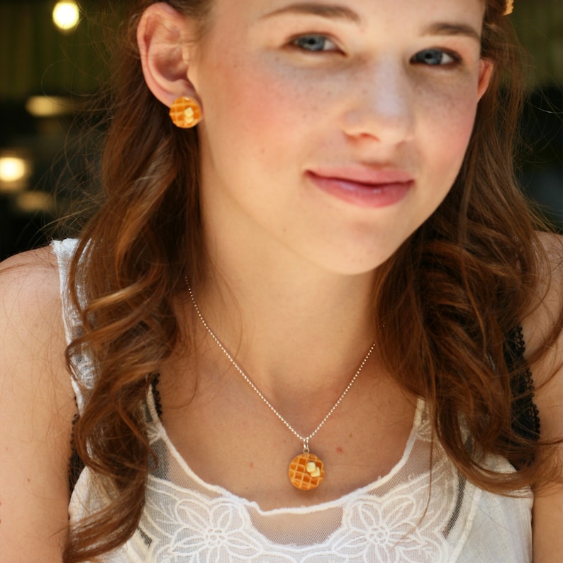 Op de afbeelding: Een vrouw draagt een wit kanten topje met een zilveren kettingketting en een wafelhanger. Ze draagt ook een paar wafel oorbellen.