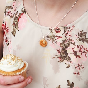 Op de afbeelding: Een zilveren ketting met een klein, gedetailleerd, bruin en geel wafelbedeltje. De ketting wordt gedragen door iemand die een blouse met bloemenprint draagt en een cupcake met witte frosting en gouden sprinkles vasthoudt.