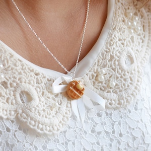 Op de afbeelding: Een zilveren ketting met een witte strik en een klein, bruin wafel-hanger. De wafel heeft een gele dot boter erop.