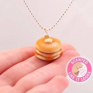 May include: A silver chain necklace with a small, three-stack pancake charm. The pancake is brown and has a dollop of white butter and a drizzle of syrup. The charm is labeled "Scented Like It Looks".