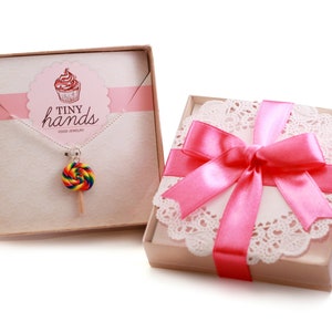 May include: A pink gift box with a white doily and a pink ribbon. The box contains a necklace with a rainbow lollipop charm. The box has the text "Tiny Hands Food Jewelry" on it.