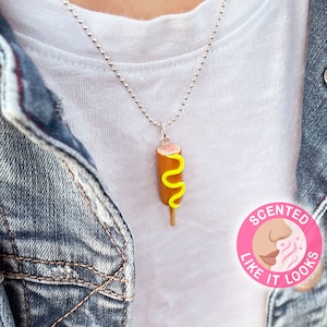 May include: A silver chain necklace with a miniature corndog pendant. The corndog is yellow and brown with a swirl of yellow mustard on top. The necklace has a tag that says "Scented Like It Looks".