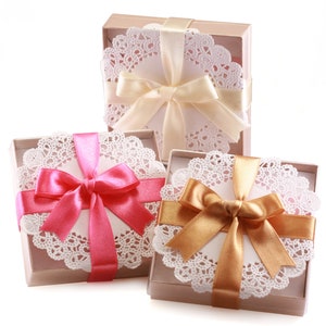 May include: Three gift boxes with white doilies and satin ribbon bows. The boxes are wrapped in brown paper and tied with pink, gold, and white ribbon.