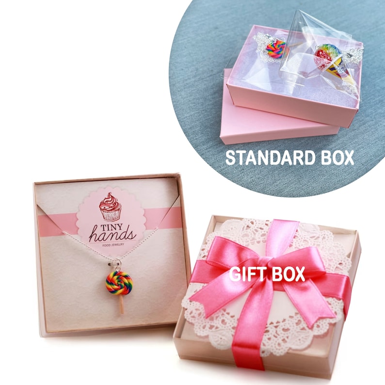 May include: Two gift boxes for jewelry. One box is a standard pink box with a clear lid. The other box is a white box with a pink ribbon and a lace doily. The box has the text "GIFT BOX" printed on it. The jewelry is a rainbow lollipop necklace with the text "TINY hands FOOD JEWELRY" on the card.