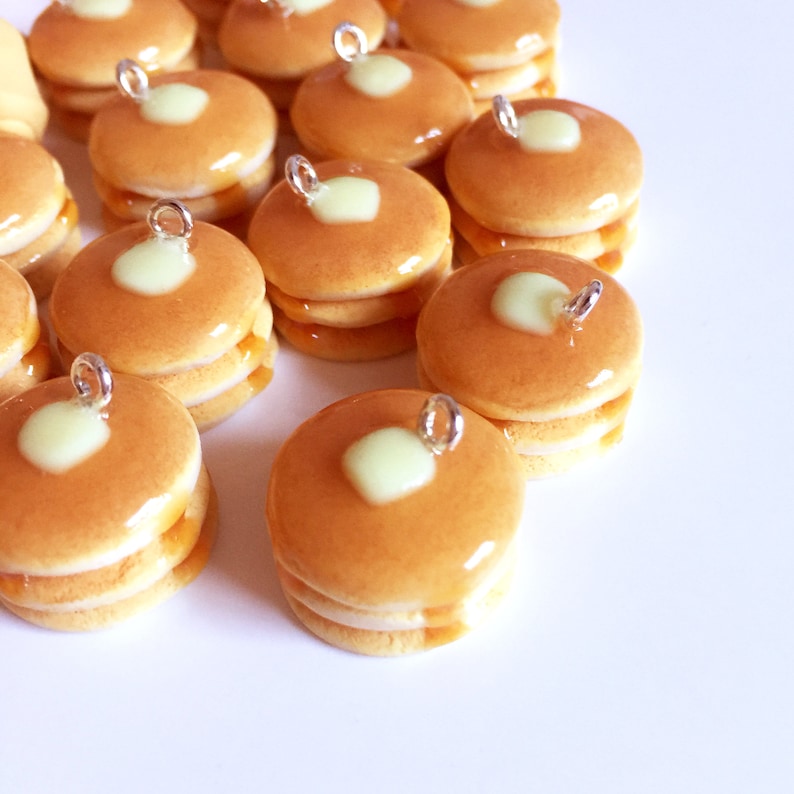 May include: A collection of miniature pancake charms with a light brown color and a light green center. The charms are stacked on top of each other and have a silver loop for hanging.