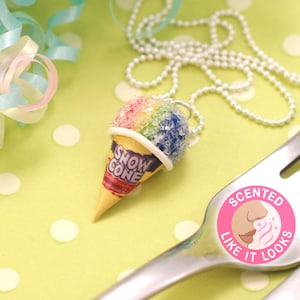 May include: A colorful snow cone charm necklace with a white chain. The charm is shaped like a snow cone with a yellow and red wrapper that says "Snow Cone". The charm is on a green and white polka dot surface.