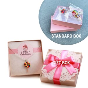 Op de afbeelding: Twee cadeaudozen voor sieraden. E&eacute;n doos is een standaard roze doos met een doorzichtig deksel. De andere doos is een witte doos met een roze lint en een kantonderzetter. De doos heeft de tekst "GIFT BOX" erop gedrukt. De sieraden zijn een regenbooglolli-ketting met de tekst "TINY hands FOOD JEWELRY" op de kaart.