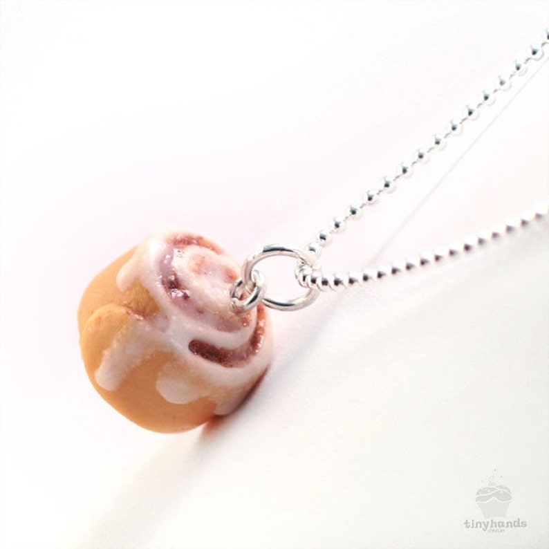 May include: A silver chain necklace with a white icing-covered cinnamon roll pendant. The roll is brown and has a swirl of cinnamon inside.