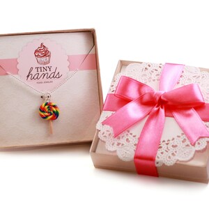 May include: A pink gift box with a white doily and a pink ribbon. The box contains a necklace with a rainbow lollipop charm. The box has the text "Tiny Hands Food Jewelry" on it.