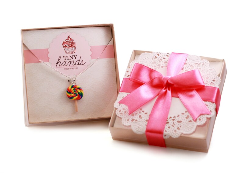 May include: A pink gift box with a white doily and a pink ribbon. The box contains a necklace with a rainbow lollipop charm. The box has the text "Tiny Hands Food Jewelry" on it.