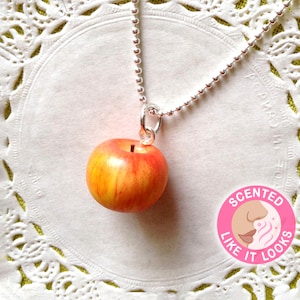 May include: A silver chain necklace with a red and yellow apple pendant. The pendant is made to look like a real apple and has a stem and leaf. The necklace is labeled "Scented Like It Looks".