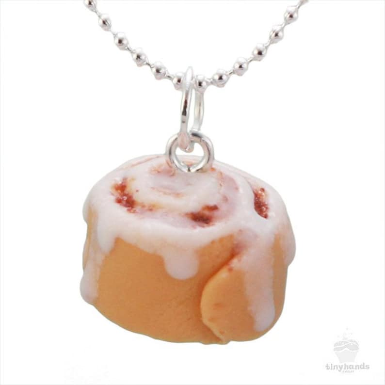 May include: A silver chain necklace with a cinnamon roll pendant. The pendant is made of polymer clay and is glazed with white icing. The cinnamon roll is a light brown color with a swirl of brown cinnamon filling.
