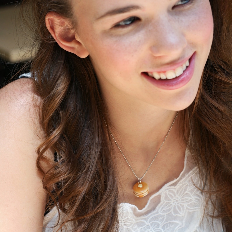 May include: A woman with long brown hair is wearing a white lace top and a silver chain necklace with a pancake charm.