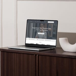 K&ouml;nnte beinhalten: Ein silberner Laptop zeigt eine Website mit dem Text "Transform Your Passion Into a Thriving Business" auf einem dunkelbraunen Schrank. Rechts vom Laptop befindet sich ein wei&szlig;es, gebogenes Dekorationsobjekt.