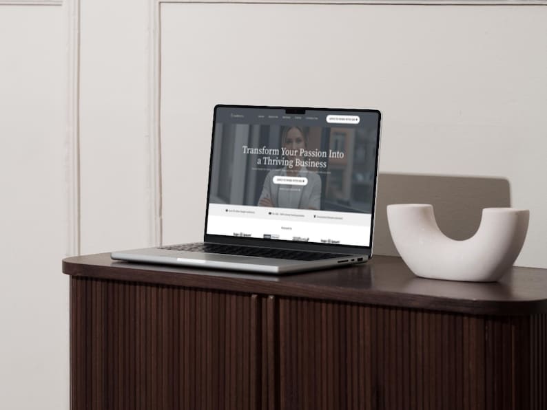 K&ouml;nnte beinhalten: Ein silberner Laptop zeigt eine Website mit dem Text "Transform Your Passion Into a Thriving Business" auf einem dunkelbraunen Schrank. Rechts vom Laptop befindet sich ein wei&szlig;es, gebogenes Dekorationsobjekt.