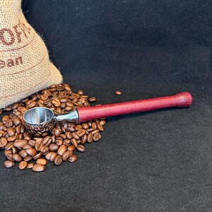 May include: A coffee scoop featuring a polished silver metal bowl and a long, cylindrical handle made of deep purple-red wood, resting atop a pile of roasted brown coffee beans next to a tan burlap sack with partial text visible.