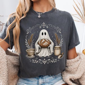May include: A charcoal gray short-sleeve t-shirt featuring a graphic print of a ghost holding a loaf of bread, surrounded by wheat stalks, a mixing bowl, and a jar, all enclosed within a decorative circular frame of swirling line art.