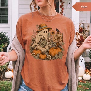 May include: A burnt orange short-sleeve t-shirt featuring a graphic of a ghost wearing a straw hat, holding a watering can and a trowel. The design includes pumpkins, sunflowers, a black bird, and a wooden sign that reads HELLO FALL Y'ALL.