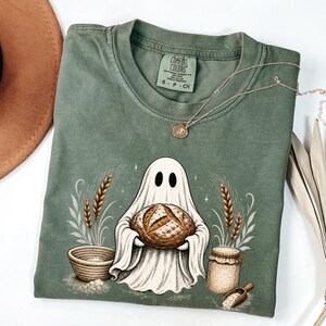 May include: A sage green short-sleeve t-shirt featuring a graphic of a white ghost holding a round loaf of bread. The design includes wheat stalks, a woven basket, a glass jar, and a small scoop. The neck label reads Comfort Colors S P CH.