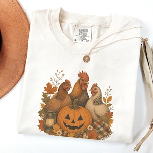 May include: A cream-colored t-shirt featuring a graphic of three chickens, a jack-o'-lantern, a lantern, and autumn leaves. The inner neck label reads Comfort Colors S P CH. A gold-toned necklace with a small circular pendant rests on the fabric.