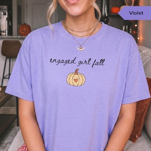 May include: A violet short-sleeve t-shirt featuring the black text "engaged girl fall" above a graphic of a light yellow pumpkin with a small orange heart in the center. The shirt has a crew neckline and a relaxed fit.
