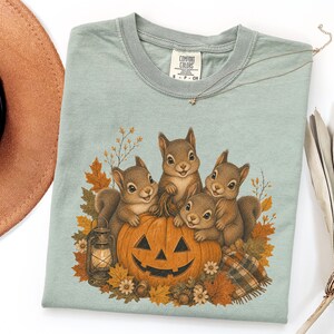 May include: A sage green t-shirt featuring a graphic of four squirrels gathered around a carved jack-o'-lantern. The design includes autumn leaves, a small lantern, and a plaid blanket. The shirt has a Comfort Colors brand label at the neck.