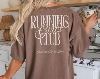 May include: A brown t-shirt with the text "RUNNING Girls CLUB" in a white, stylized font on the back. Below the main text is the phrase "Girls Who Run the World" in a smaller, cursive font. The shirt is a solid color.