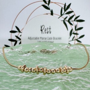 May include: A gold-coloured adjustable Morse code bracelet with the word "Rest" on the card behind it. The bracelet features small, round beads and cylindrical accents. The bracelet is displayed on a textured, light green surface.