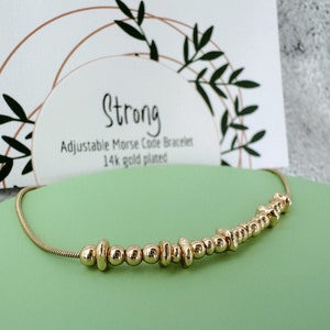 May include: Gold-toned adjustable bracelet with beads spelling out the word "strong" in Morse code. The bracelet is on a green surface with a card that says "Strong Adjustable Morse Code Bracelet 14k gold plated".