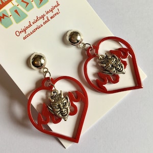 Könnte beinhalten: Rote herzförmige Ohrringe mit einem silbernen Teufelskopf-Anhänger. Die Ohrringe befinden sich auf einer weißen Karte mit dem Text "Mid Century Missy Original vintage inspired accessories and more!"